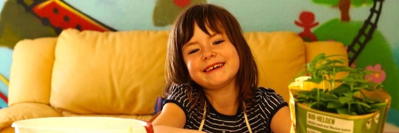 Ein Mädchen sitzt an einem Tisch und knetet Teig. Sie schaut lachend in die Kamera. Hinter ihr ist eine bunt angemalte Wand.